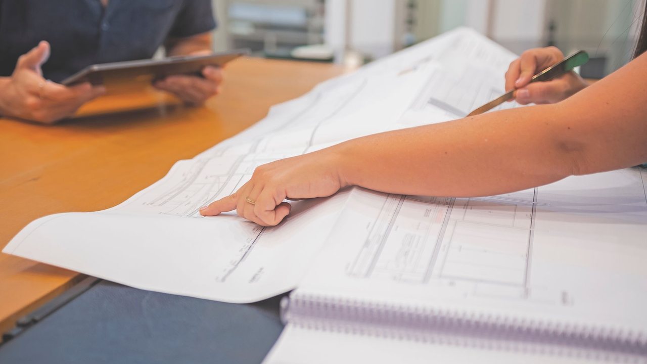 Manos señalando un plano sobre una mesa junto a una tableta