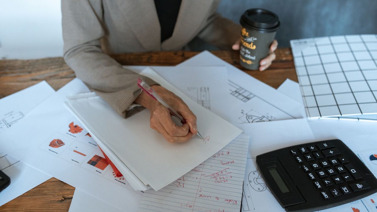 Persona escribiendo en un cuaderno junto a documentos y una calculadora